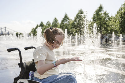 Cheerful girl with disability sitting on wheelchair playing by fountain