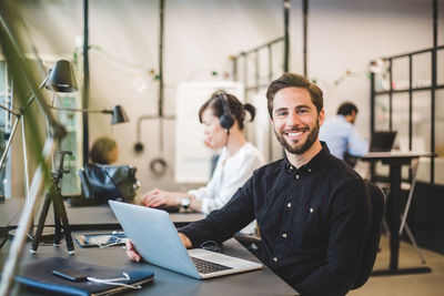 Portrait of confident businessman with laptop at desk in creative office