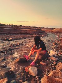 Woman sitting on rock at shore against sky during sunset