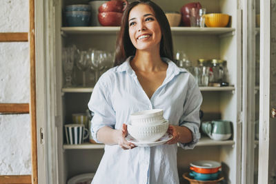 Smiling young woman holding crockeries while standing at home