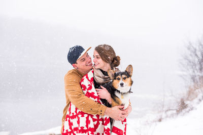 Young couple smile and huddle during snowfall with dog