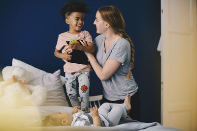 Mother holding son while standing by toddler on bed at home