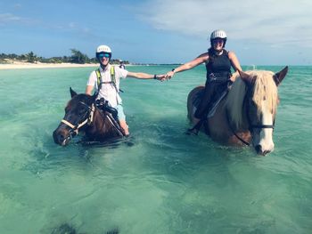 Friends holding hands while sitting on horses in sea