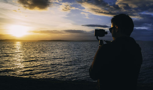 Rear view of silhouette man photographing sea against sky during sunset