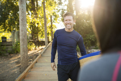 Portrait of man carrying paddleboard while standing with woman on boardwalk