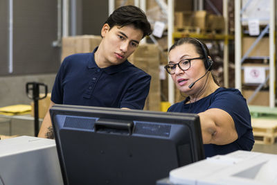 Confident mature female customer service representative discussing with coworker over computer monitor at distribution w