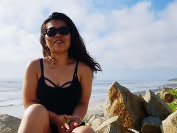 Smiling woman wearing sunglasses while sitting at beach against sky