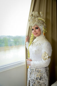 Portrait of woman wearing wedding dress standing by window