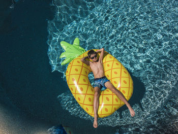 High angle view of shirtless boy with hands behind head relaxing on inflatable ring in swimming pool