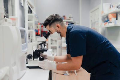 Side view of crop anonymous male medic in uniform and mask using microscope while working in lab