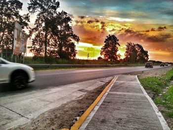 Road by trees against sky during sunset