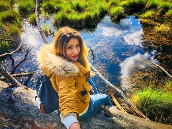 Portrait of smiling woman sitting on fallen tree by lake