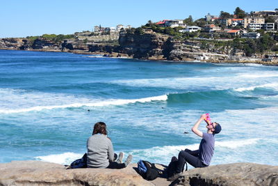 Friends sitting on rock by sea against clear sky