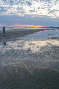 Full length of man holding camera at beach standing against sky during sunset