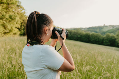 Young woman standing on meadow, using camera and taking photos at golden hour