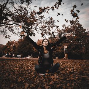Smiling young woman with arms raised throwing autumn leaves while sitting on field in park