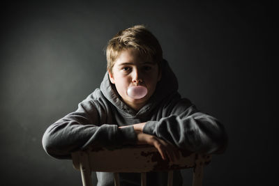 Teen boy in hoodie sitting on a chair in dark room blowing a bubble.