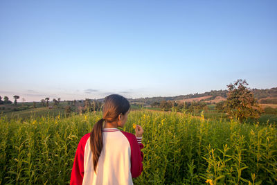 Rear view of young woman holding flower while standing amidst plants on field against sky