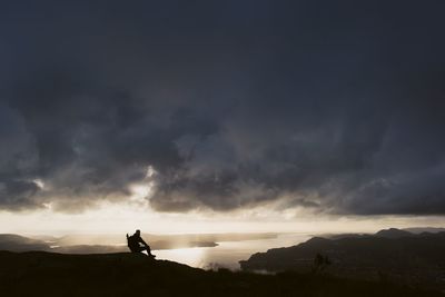 Silhouette man sitting on mountain against stormy clouds during sunset