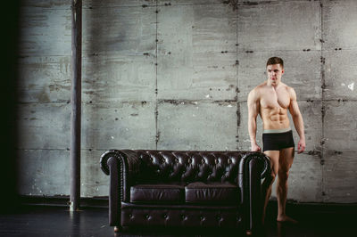 Shirtless muscular man standing by armchair against wall