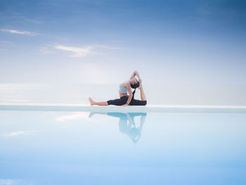 Woman practicing yoga on infinity pool by sea against cloudy sky