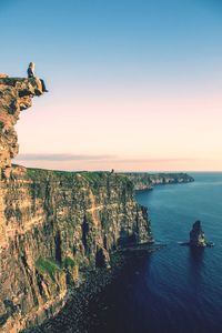 Woman sitting on cliff by sea against sky during sunset