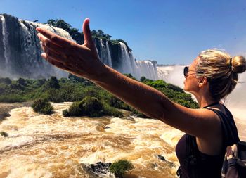 Side view of woman with arms outstretched standing against waterfall