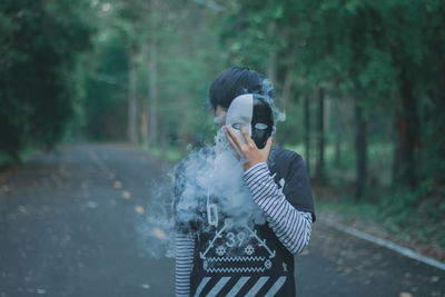 Man holding mask with smoke over face while standing against trees in forest