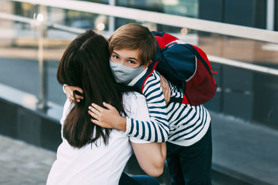 A mother hugs her young son goodbye near a school or kindergarten. 