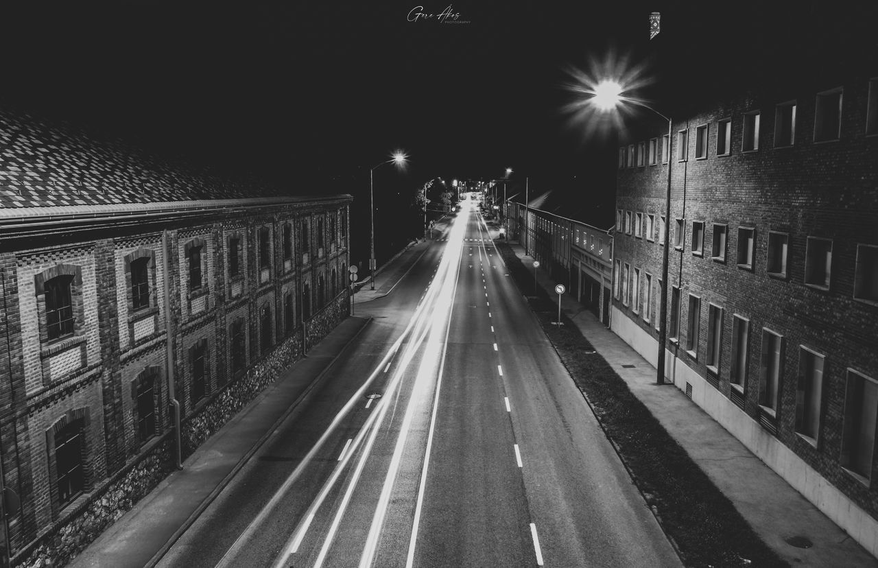 transportation, night, darkness, black and white, architecture, illuminated, road, city, monochrome, street, monochrome photography, built structure, mode of transportation, light, motion, the way forward, street light, building exterior, lane, no people, infrastructure, lighting equipment, diminishing perspective, outdoors, black, sky, nature, sign, travel, long exposure, vanishing point, city life, symbol, city street, light trail, white, line