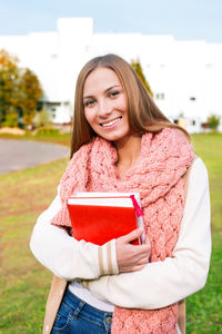 Portrait of smiling young woman holding books while standing on field