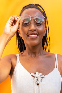 Positive african american female with braided hair touching glasses and looking at camera on yellow background in barcelona in summer