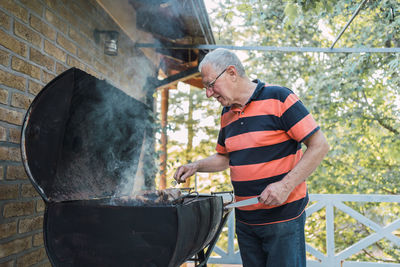 Positive senior man in casual clothes and eyeglasses frying meat on grill standing on terrace of brick house during vacation in countryside