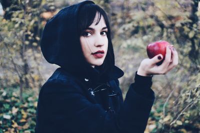 Portrait of young woman holding red apple while standing at park