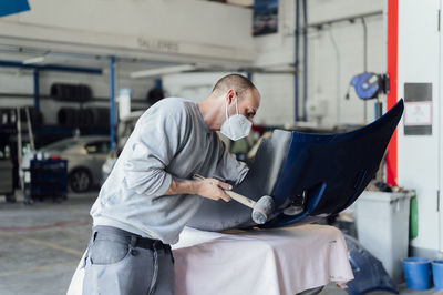 Repairman wearing protective face mask hammering on car body in workshop