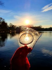 Close-up of hand holding glass against lake during sunset