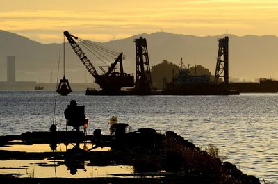 Silhouette cranes at harbor against sky during sunset