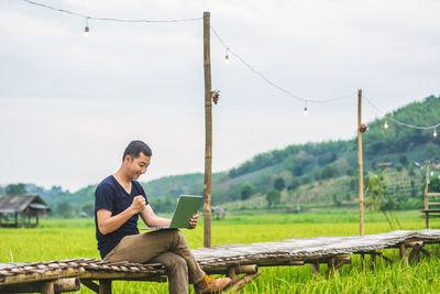 Man using laptop while sitting on boardwalk at rice paddy