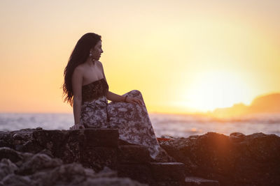 Woman sitting on rock against sky during sunset