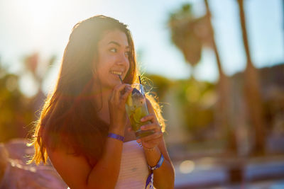 Smiling young woman looking away while drinking juice by swimming pool