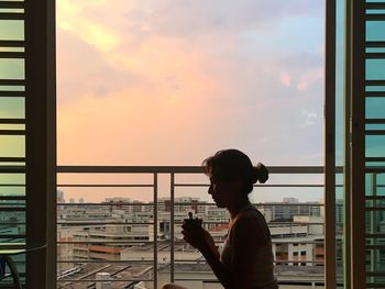 Side view of young woman looking through window at sunset