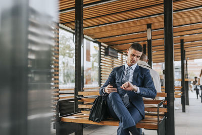 Businessman checking time while sitting on bench at bus stop