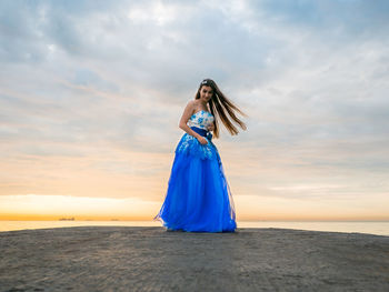 Young woman in dress standing on pier against cloudy sky during sunset