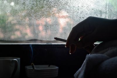 Cropped hand of woman holding cigarette by wet window at home