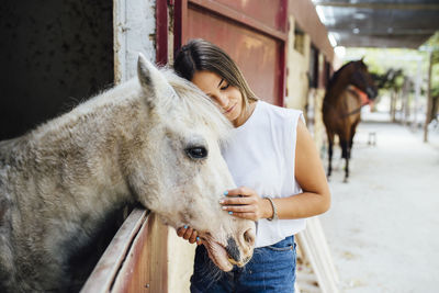 Young woman stroking pony standing in stable