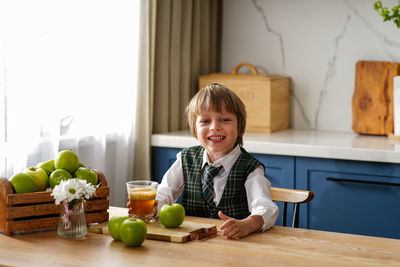 Thoughtful schoolboy is having breakfast with fresh juice and apple waiting for the school bus