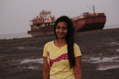 Portrait of young woman standing against old ship at beach