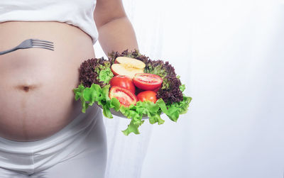 Midsection of pregnant woman holding salad bowl against white background