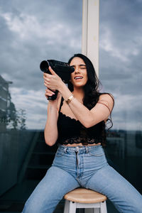 Portrait of young woman holding camera while sitting against sky