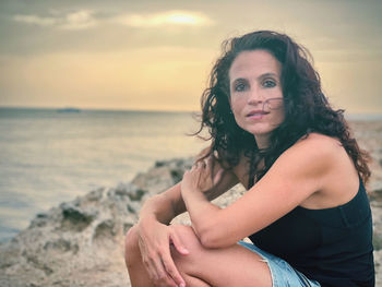 Portrait of woman sitting on beach against sky during sunset